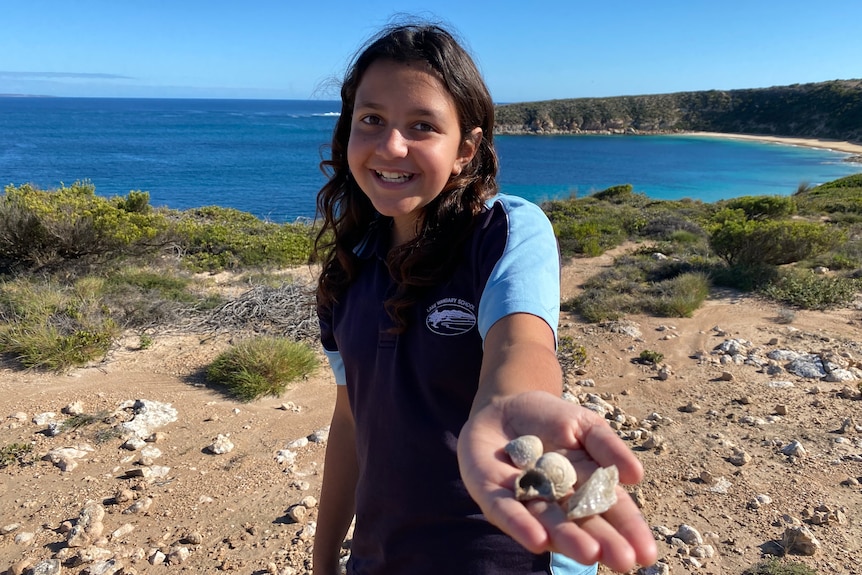 Young Indigenous girl smiling holding a hand forward with shells and a rock toward camera in front of coastal bay scene