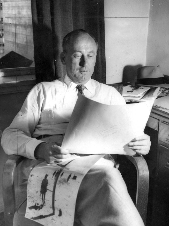 A black and white photo of a man in a white shirt and tie reading papers in an office.