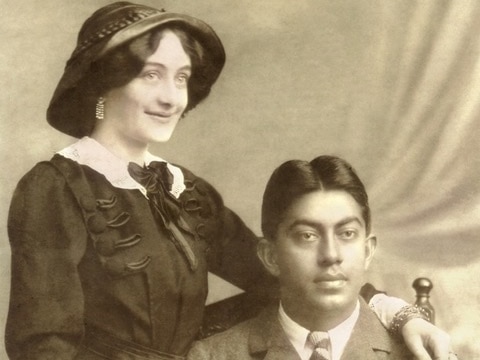 A sepia toned photo of a woman standing with her arm around an Indian man in a suit.