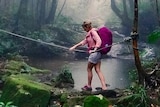 Woman in pink wearing backpack walks across creek, holding rope, in rainforest