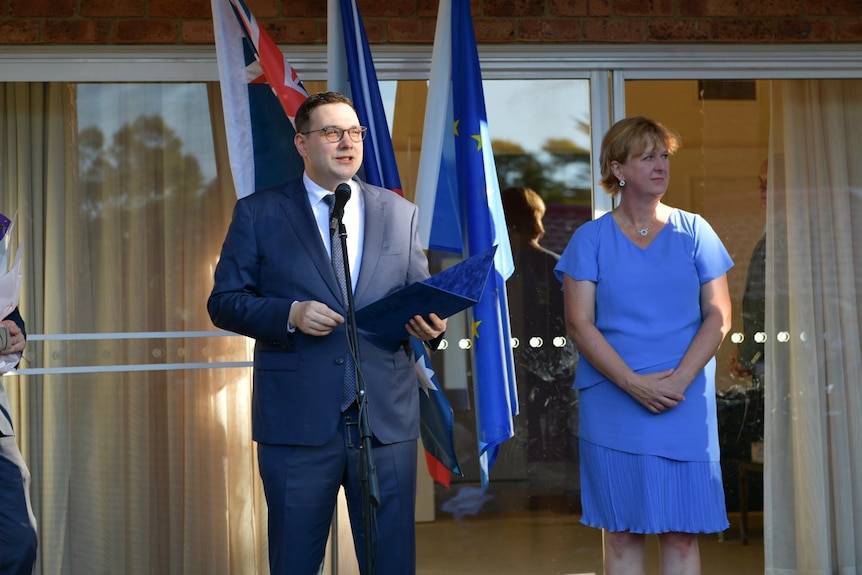 An agreeable-looking young man with glasses gives a speech from notes in a suit outsidewith the Australian, Czech and Eu flags