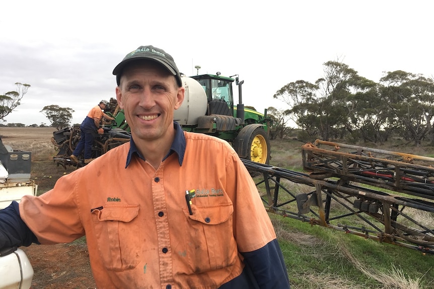 A man in an orange work shirt and a cap stands in front of a tractor.