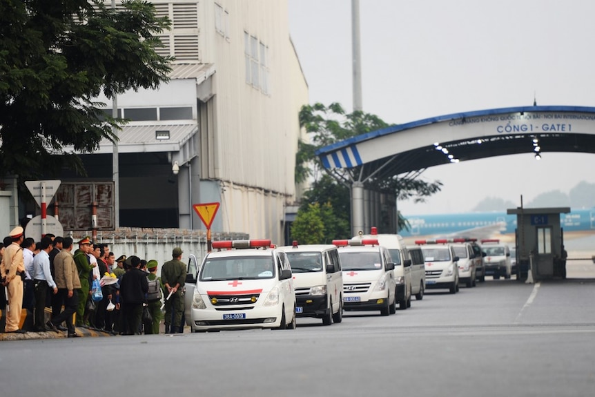 Looking across a wide street, you view a row of white ambulances parked in front of an airport gate.