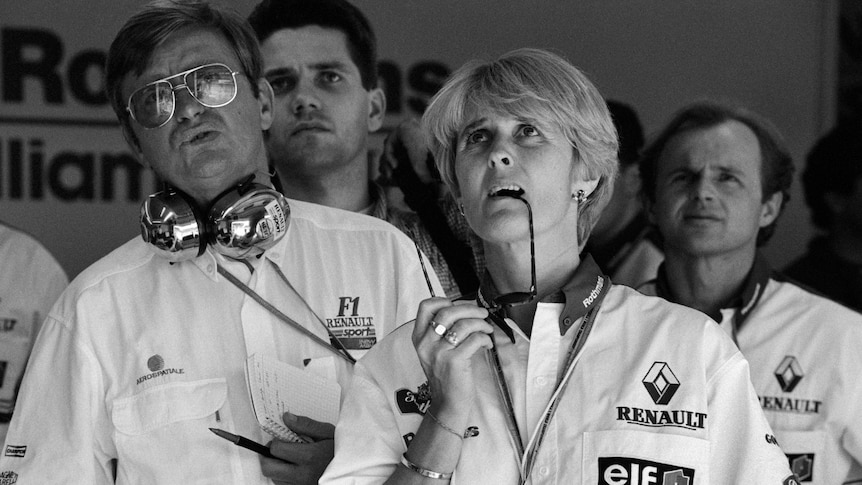 Men and women in an F1 garage look on with concern, with one woman biting her sunglasses