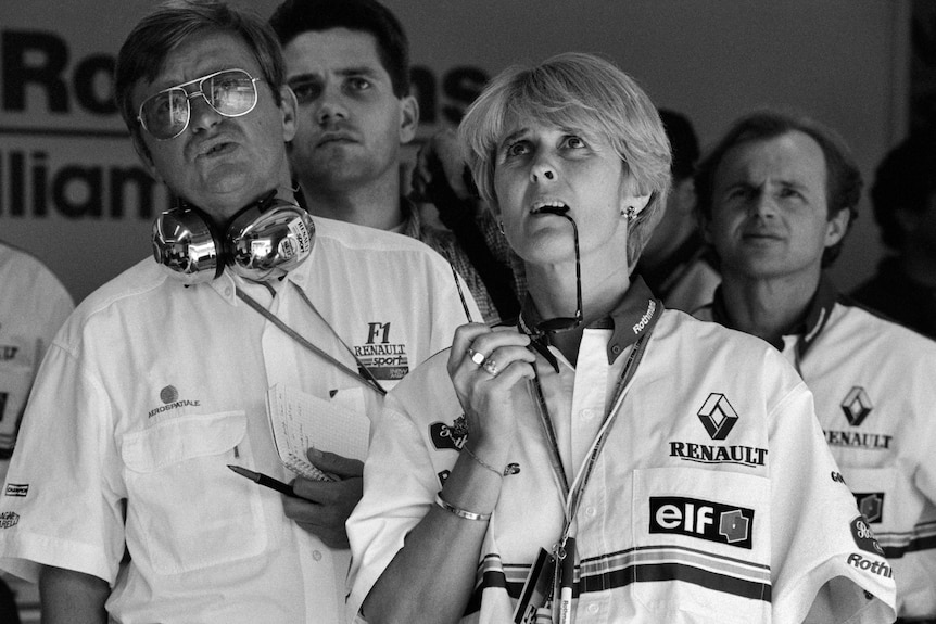Men and women in an F1 garage look on with concern, with one woman biting her sunglasses