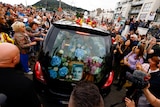 A crowd gathering around a hearse with a picture of Sinead O'Connor on display. 