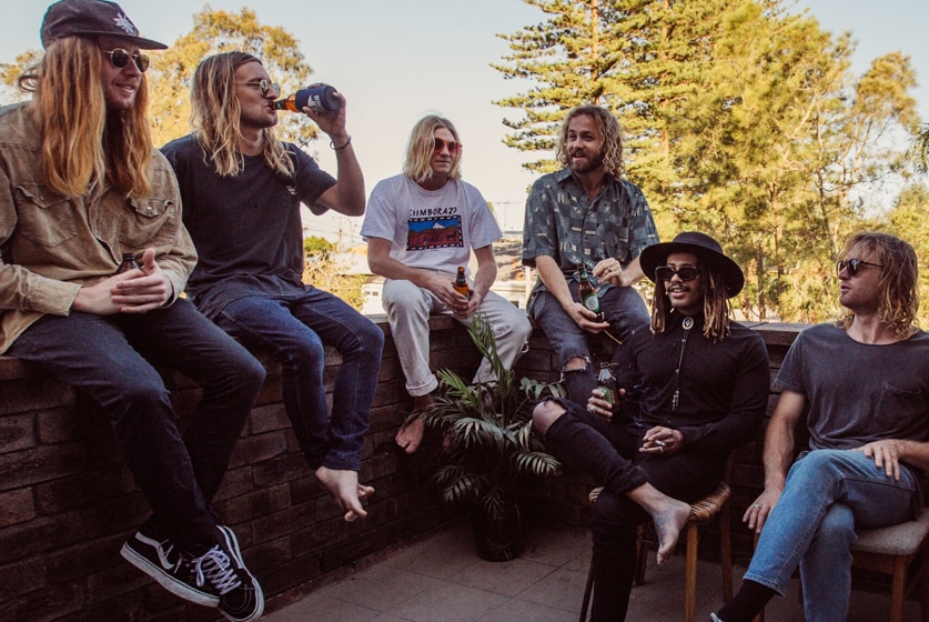 Six young men sit on a low wall and chairs with tall trees and blue sky in the background.