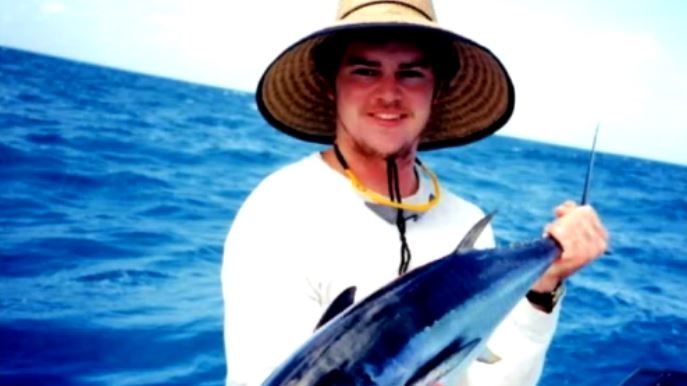 A man in a wide hat standing on a boat, holding a fish, with the sea behind him