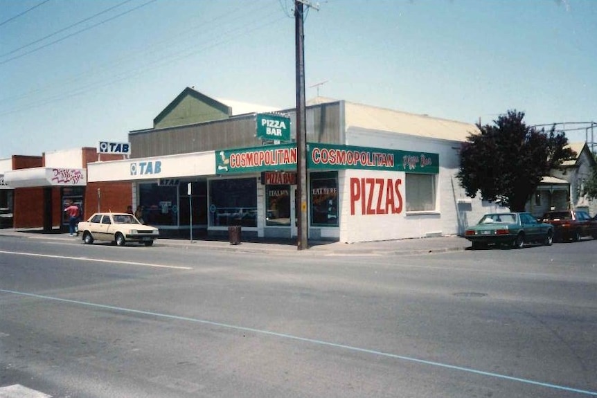 A vintage photo of a corner shop painted in red, green and white with signs reading 'Cosmopolitan Pizza Bar'.