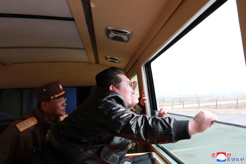 North Korean leader Kim Jong Un and two men in military uniforms look out a window and into the sky.