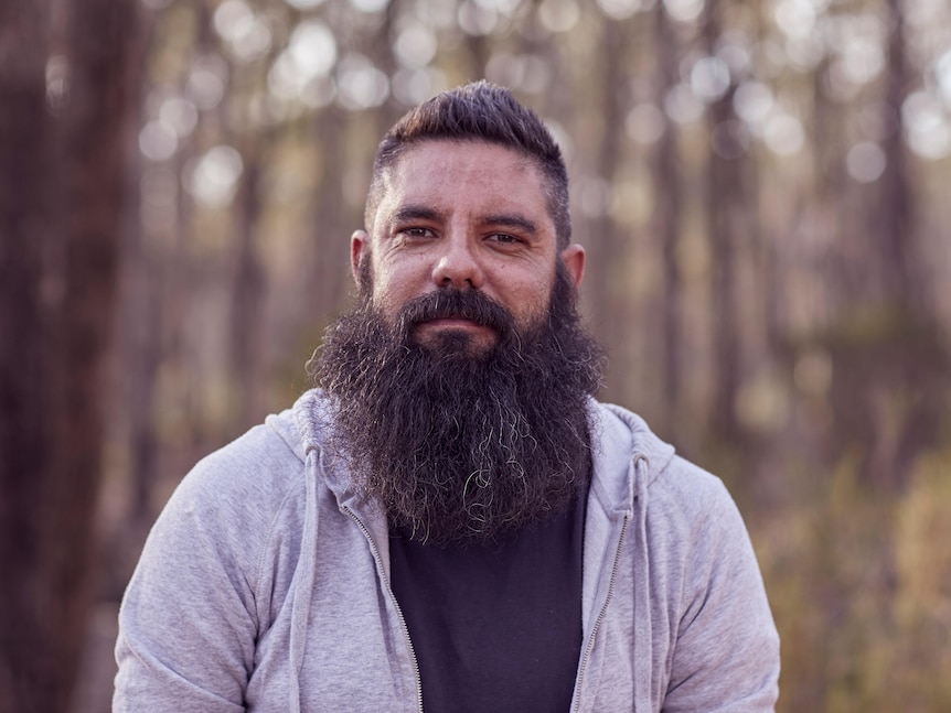 Trent Nelson poses for a photo in a national park. He has a bushy beard and is wearing a grey jacket.