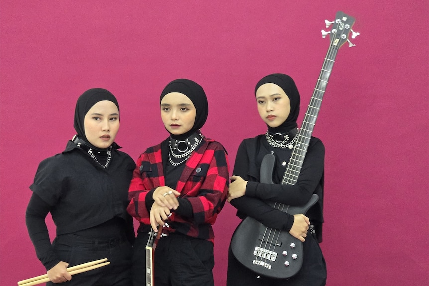 three women in black hijabs, tops and pants and chain necklaces carry their musical instruments, drumsticks, guitar and bass