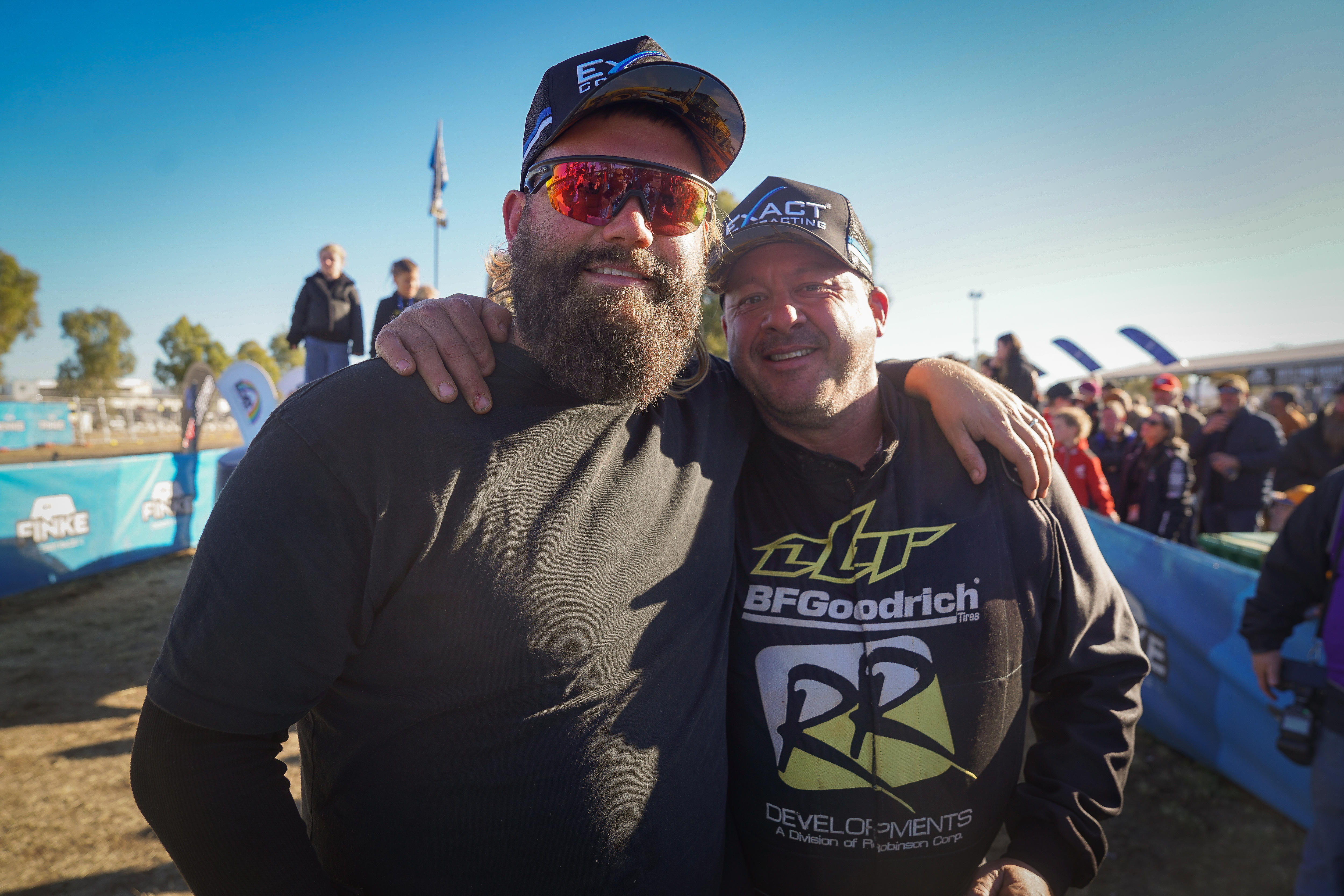 Two men standing arm-in-arm and smiling, beside a car racing track.