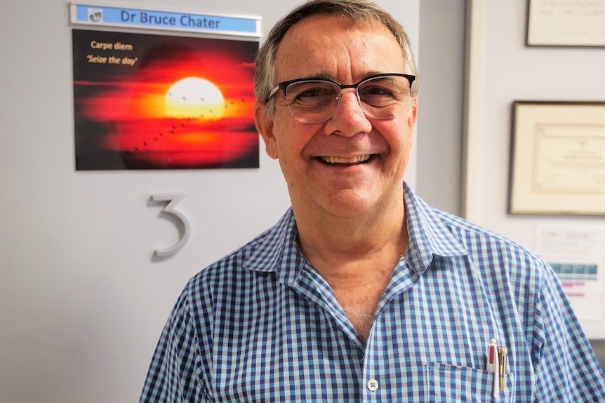 Dr Bruce Chater standing in front of his doctor's office door, smiling, wearing colourful button up and glasses.