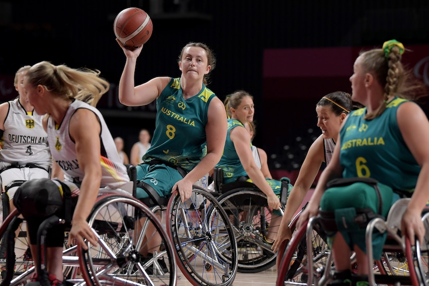 A woman in a wheelchair wearing a green and gold Australia singlet holds a basketball and prepares to pass to a teammate