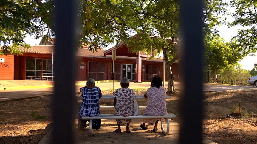 Three women sit at a table and chairs outside a brick hospital building. The photo is taken between the bars of the fence.