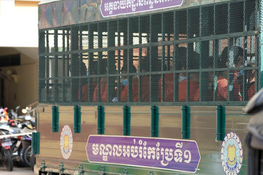 People in orange prison uniforms sit inside a cage on the back of a truck. 
