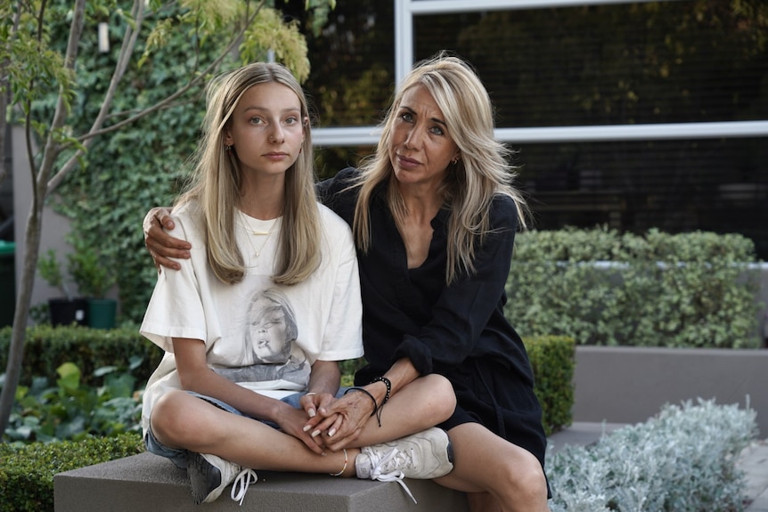 A teenage girl in a white t-shirt and her mother in a black dress sit together outside.