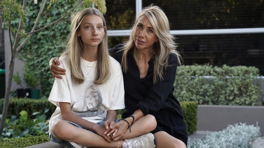 A teenage girl in a white t-shirt and her mother in a black dress sit together outside looking serious.
