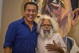 Anh Do and Uncle Jack Charles stand together smiling in front of a large portrait.