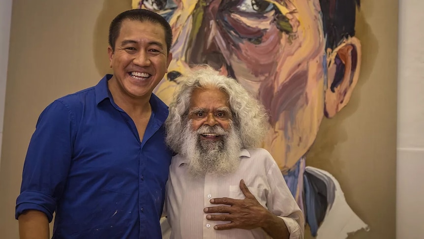 Anh Do and Uncle Jack Charles stand together smiling in front of a large portrait.