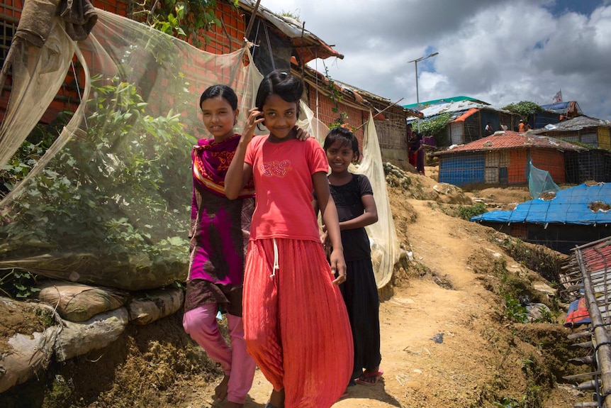 Three smiling girls walk down a muddy road in the camp