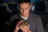 A teenage boy in a darkened bedroom holding and patting a pet tortoise.