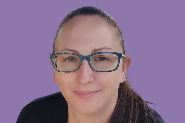 A woman with slicked back brown hair wearing dark blue framed glasses on a solid purple background