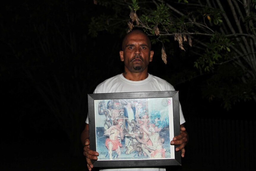 A man stands outdoors in the dark, holding a framed photo of an Aboriginal dance group.