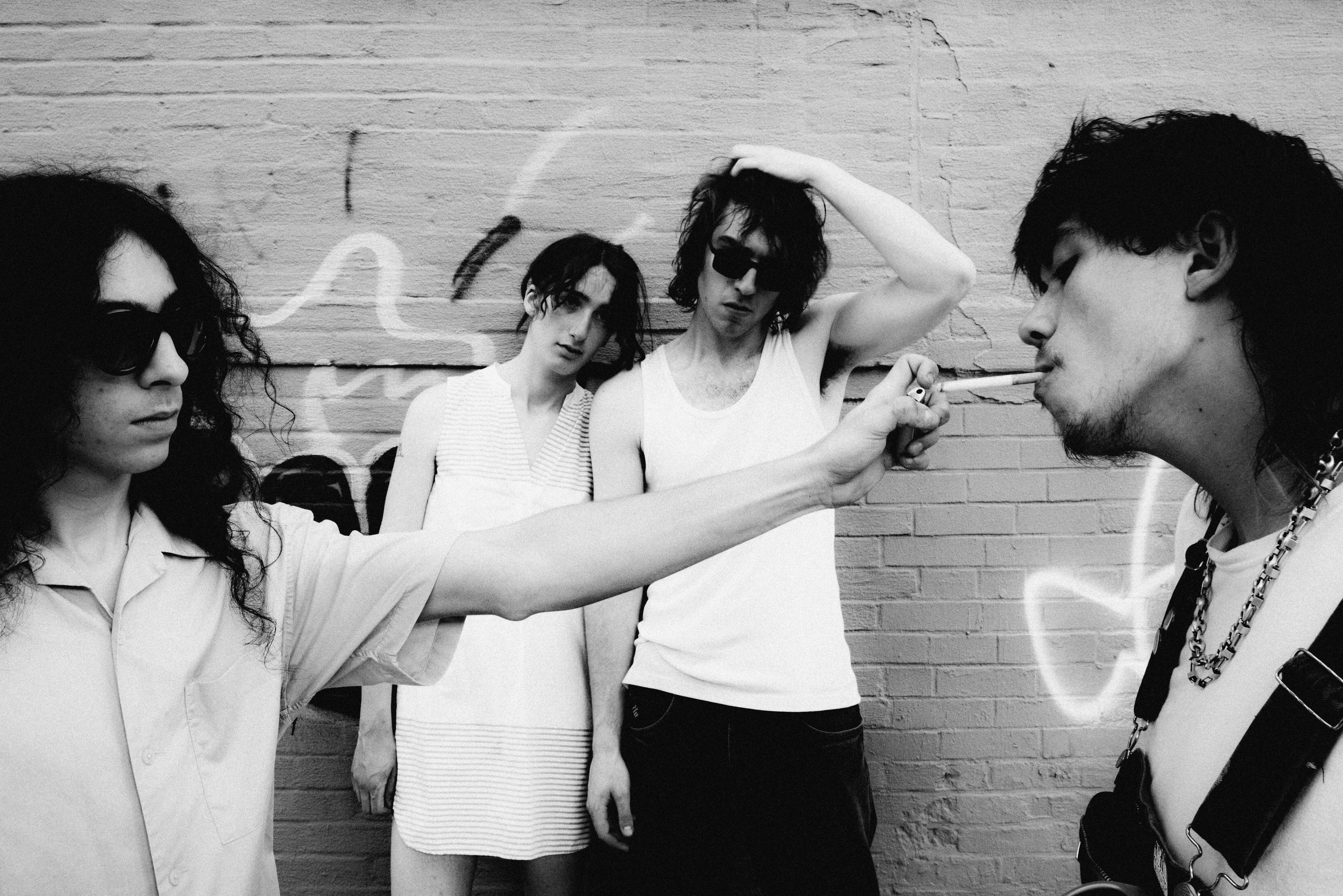 Black and white photo of three men and a woman against a concrete wall, one is lighting a cigarette for another