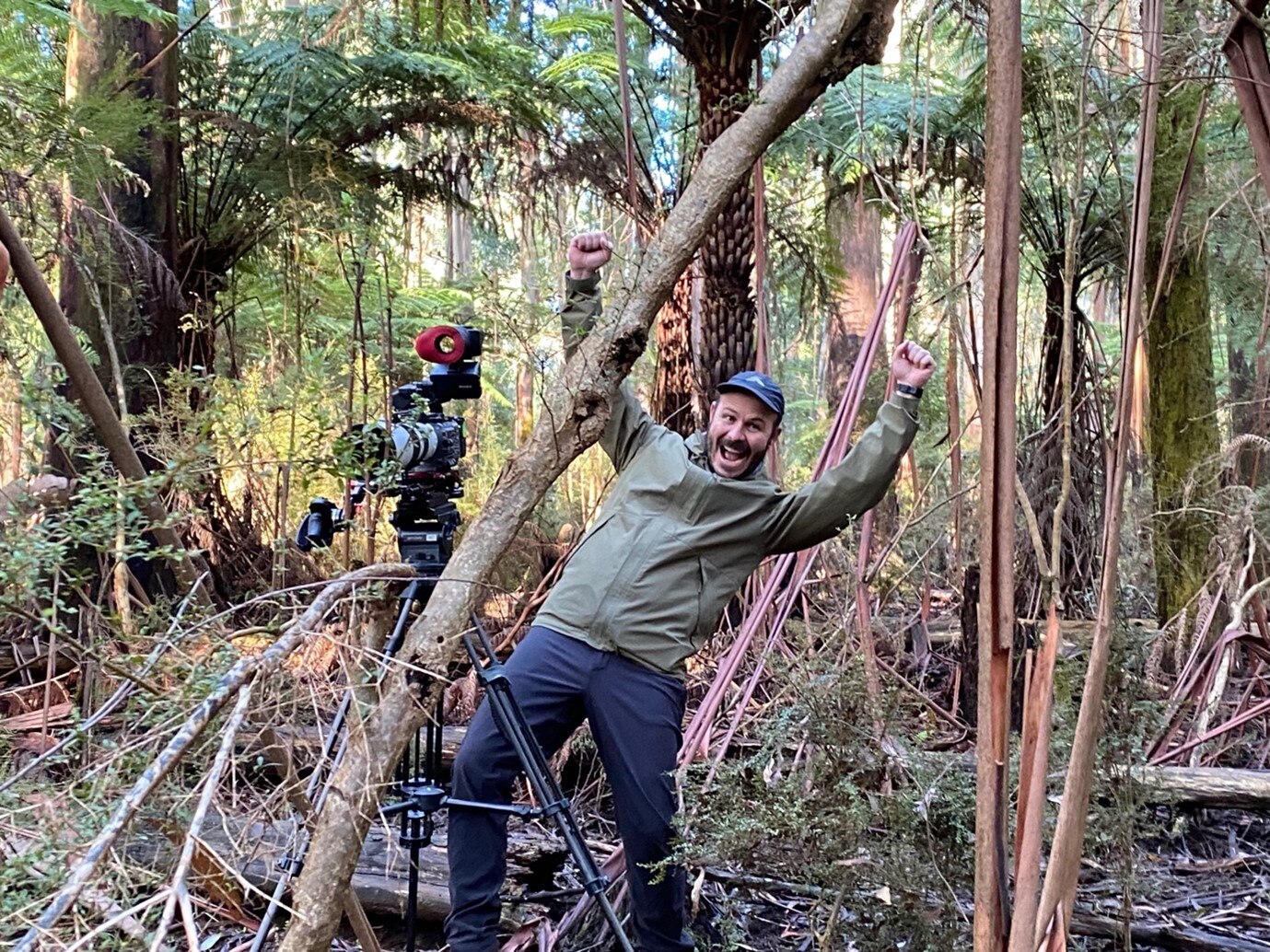 Man standing next to camera in bush with arms up as though cheering and big smile on face.