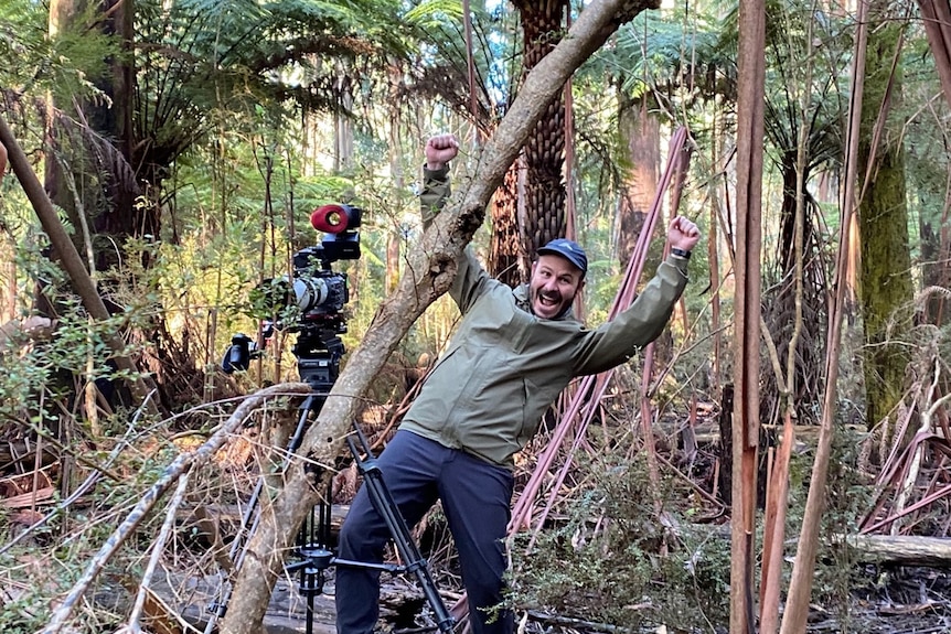 Man standing next to camera in bush with arms up as though cheering and big smile on face.