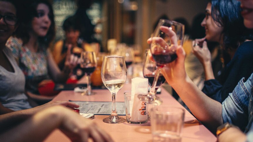 Women talking and drinking wine at a long table in a low lit bar.