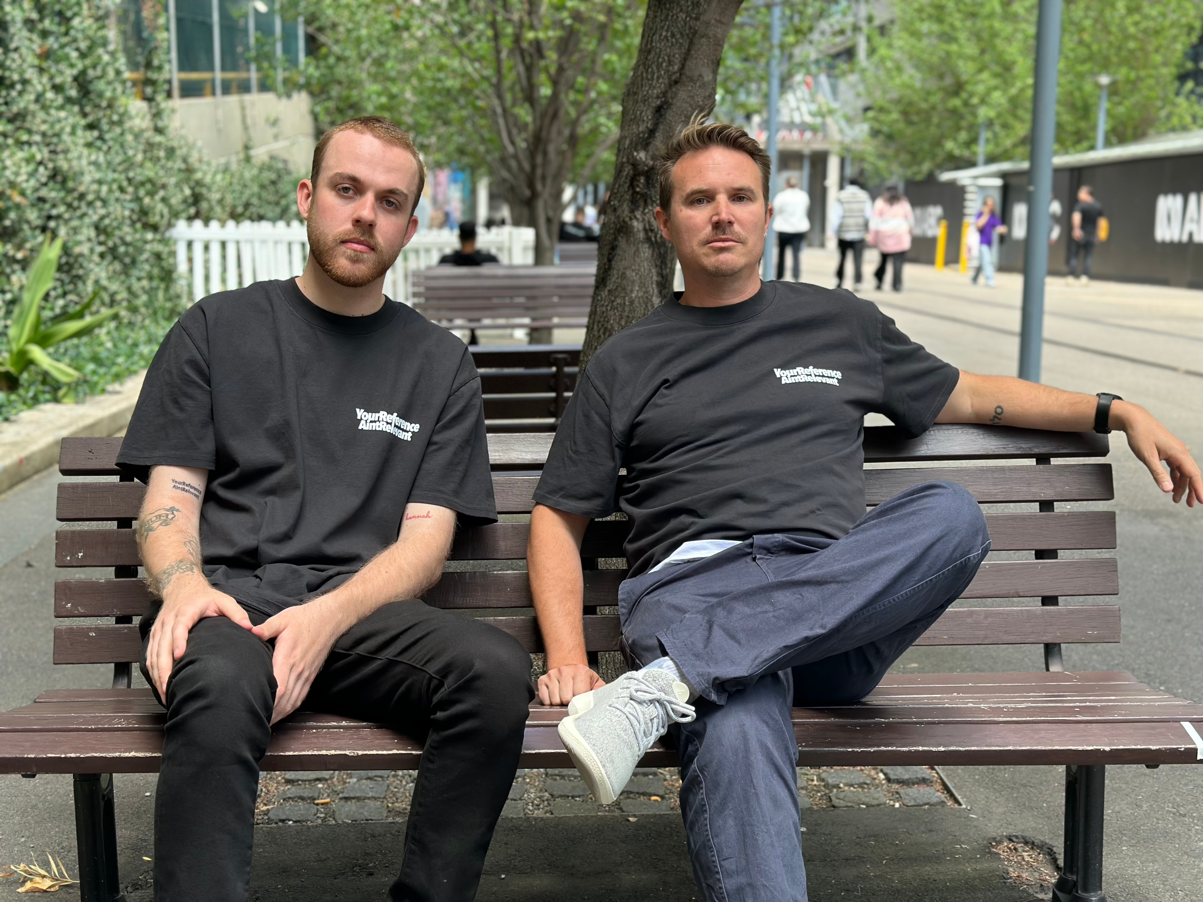 Two men sit on an outdoor bench looking serious.