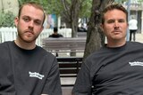 Two men sit on an outdoor bench looking serious.