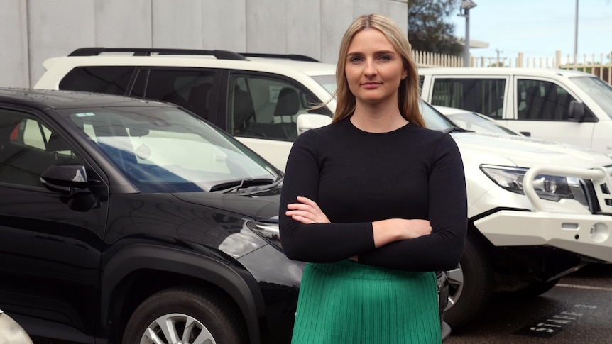 ABC reporter Keely Johnson stands with her arms crossed in front of cars in a carpark.