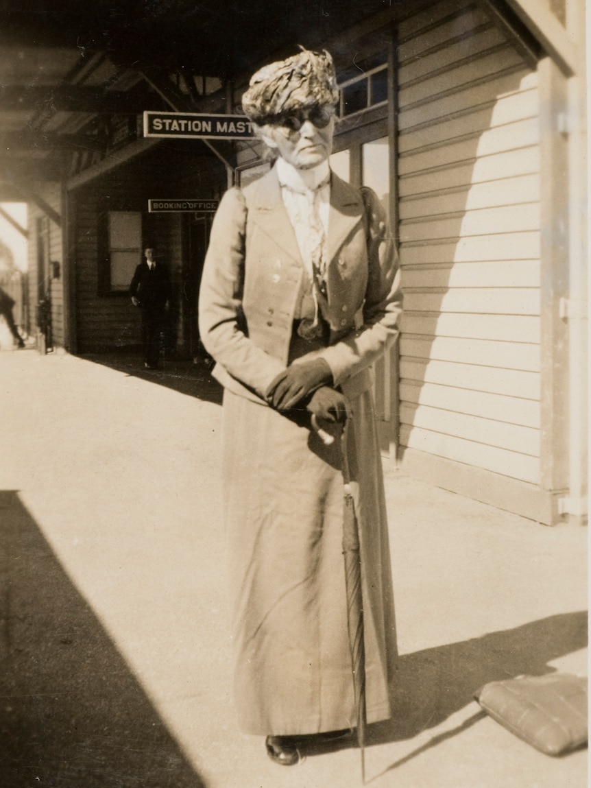 A woman dressed in a long skirt and jacket, wearing a hat and glasses and holding an umbrella, stands on a railway platform.