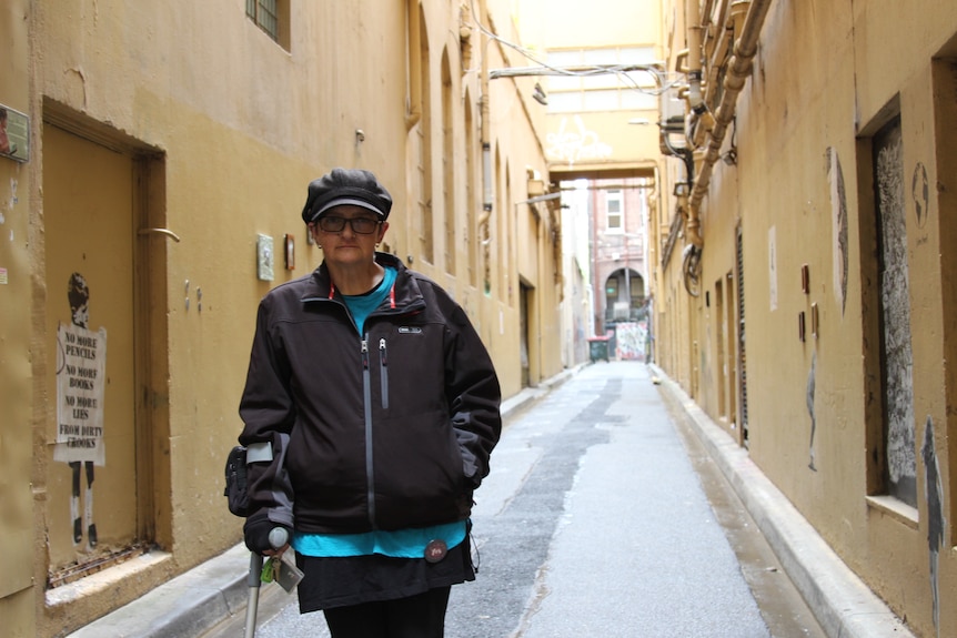 A woman who wears a black cap, glasses, jacket, blue tee, leans on a crutch, stands in a beige laneway.