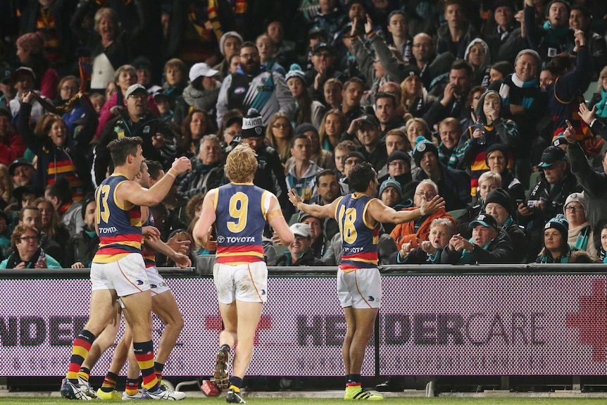 An Indigenous AFL player is surrounded by teammates as he stares at angry fans of the opposing team during a game.