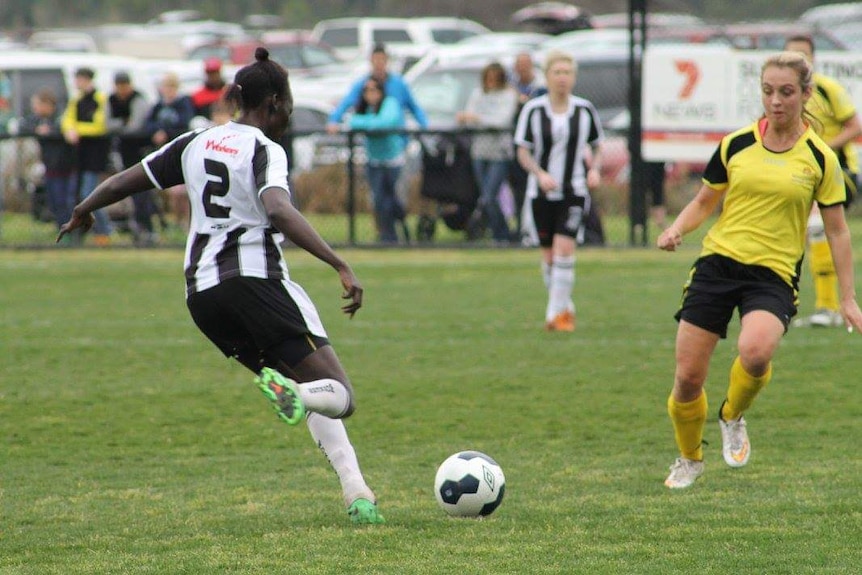 A woman is kicking a football on an oval, with another women defending her.