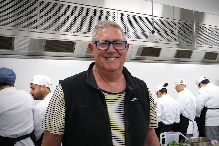 A man wearing a jacket and glasses smiles while standing in a commercial kitchen