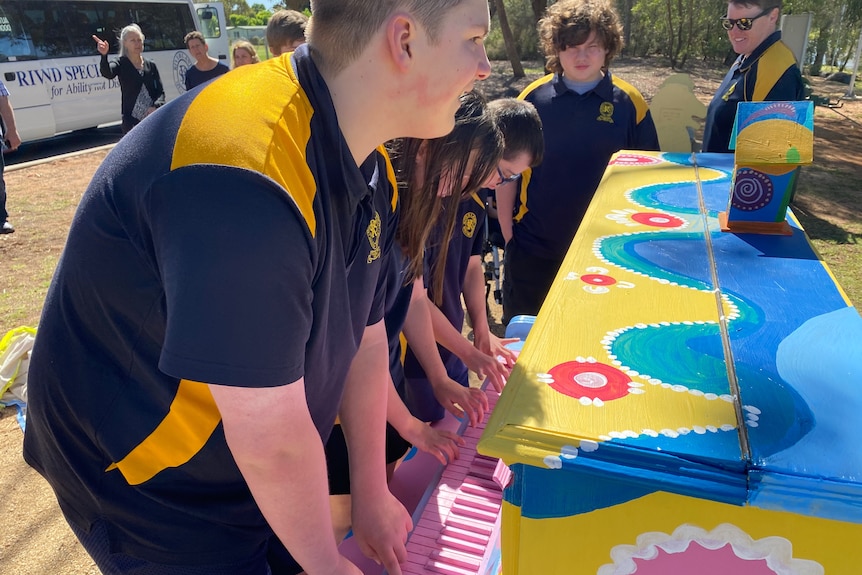 A group of children stand at a brightly-coloured piano and play it together 