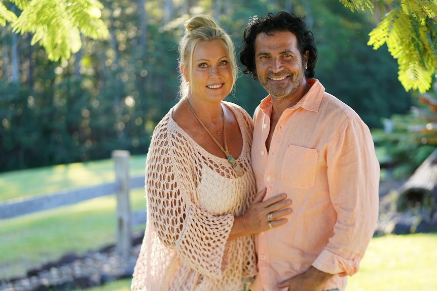 A portrait of a woman wearing a blush pink shawl standing next to husband outdoors who wears a pastel orange button-up shirt