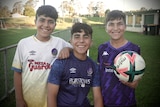 Three smiling young brothers sitting by the side of a soccer pitch.