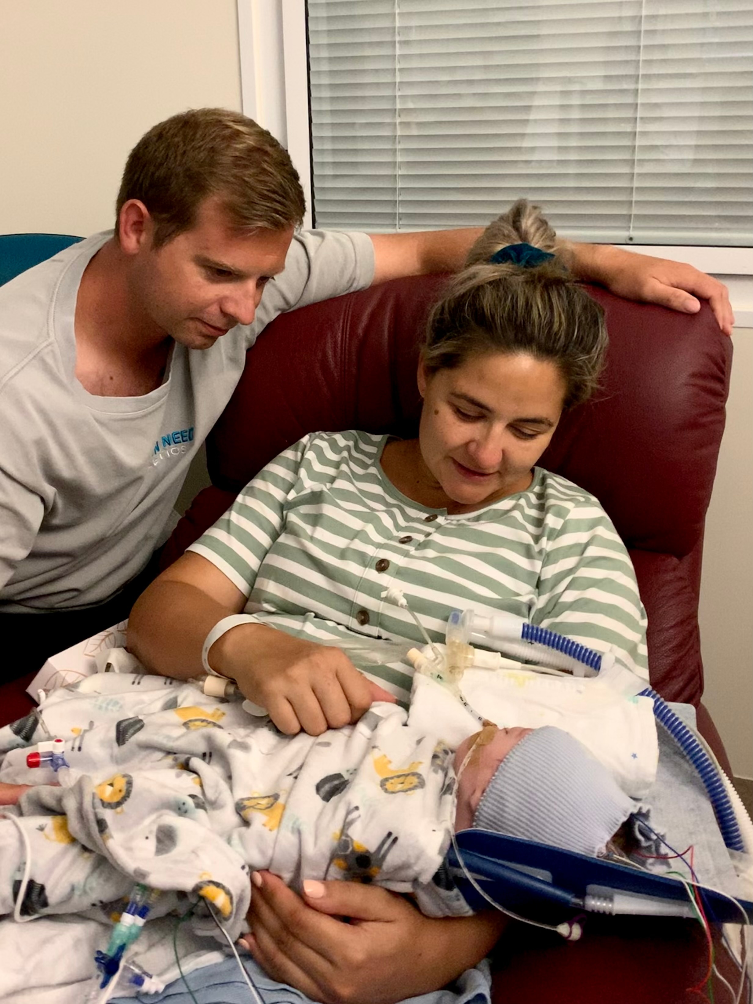 A man looks down at a newborn baby lying on the lap of a woman sitting in an armchair in a hospital.