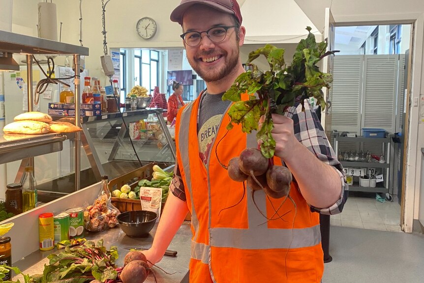 A man in a cap and hi-vis vest holds a bunch of beetroots in a kitchen