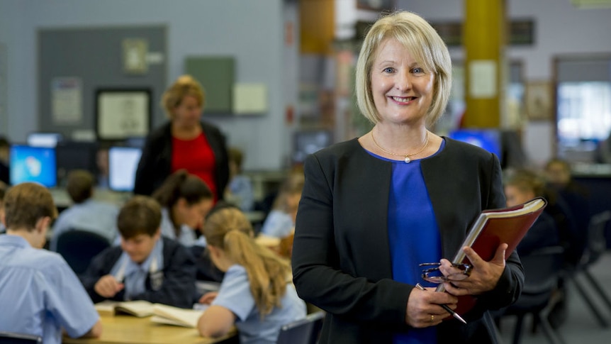 A woman with blonde hair holding a folder, with primary school students in class in the background.