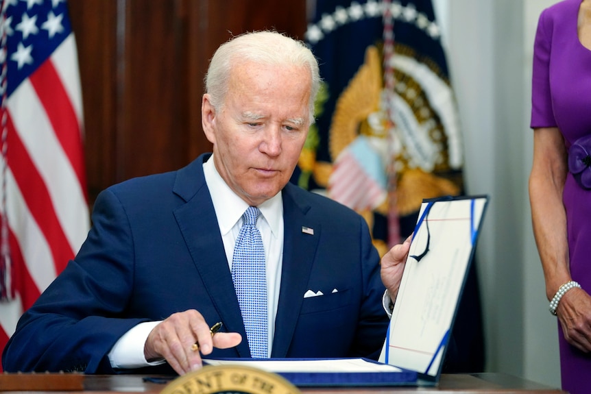 President Joe Biden sitting and holding open the file to sign.