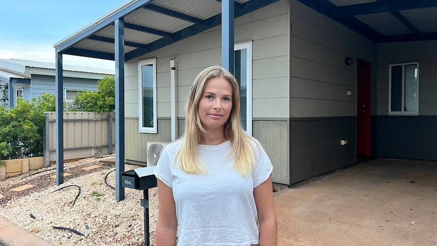 A woman with blonde hair wearing a white t-shirt stands outside a house