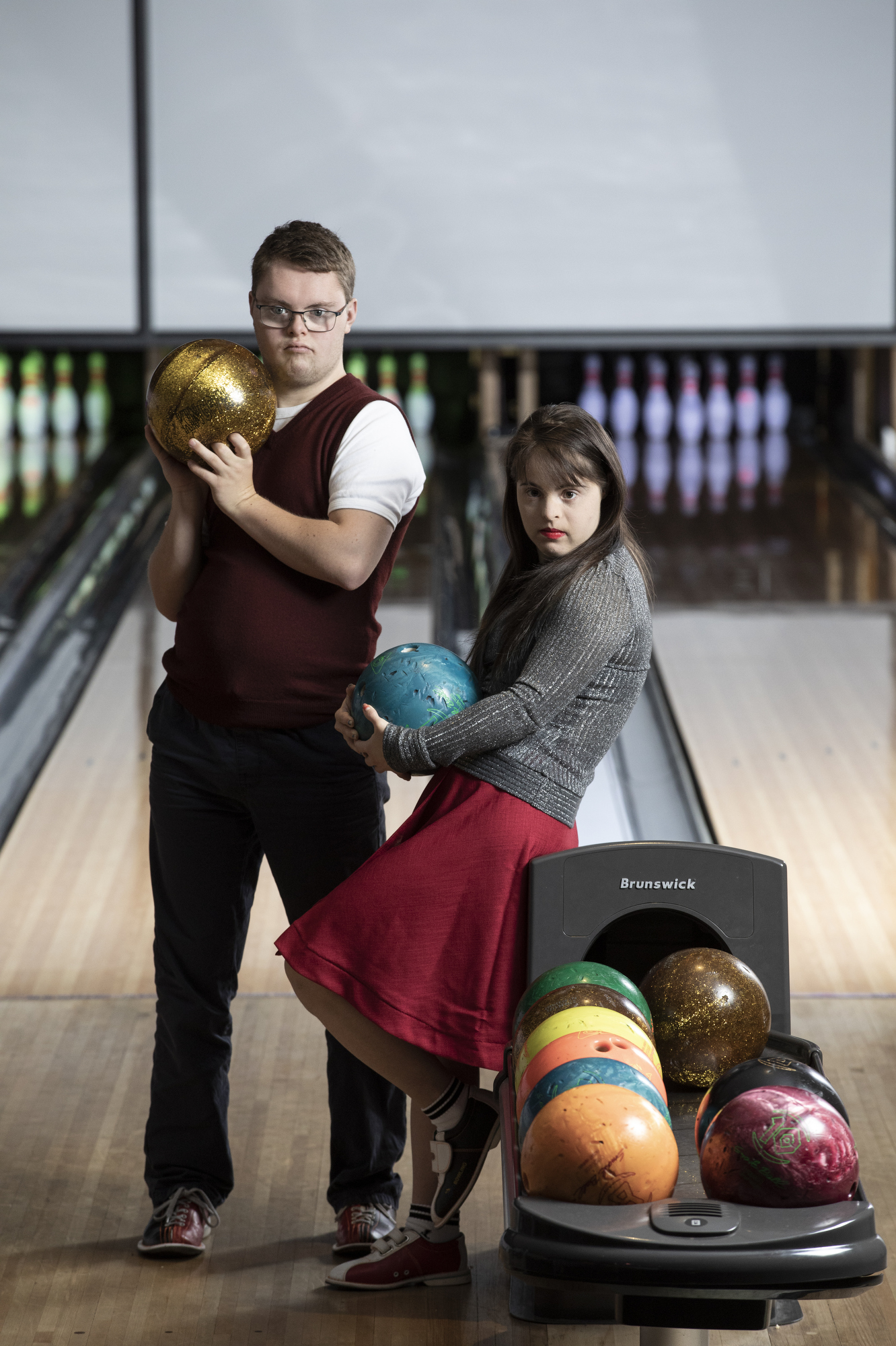 A blonde white man in a vest and pants and a brunette white girl in silver top and red skirt hold bowling balls in an alley.